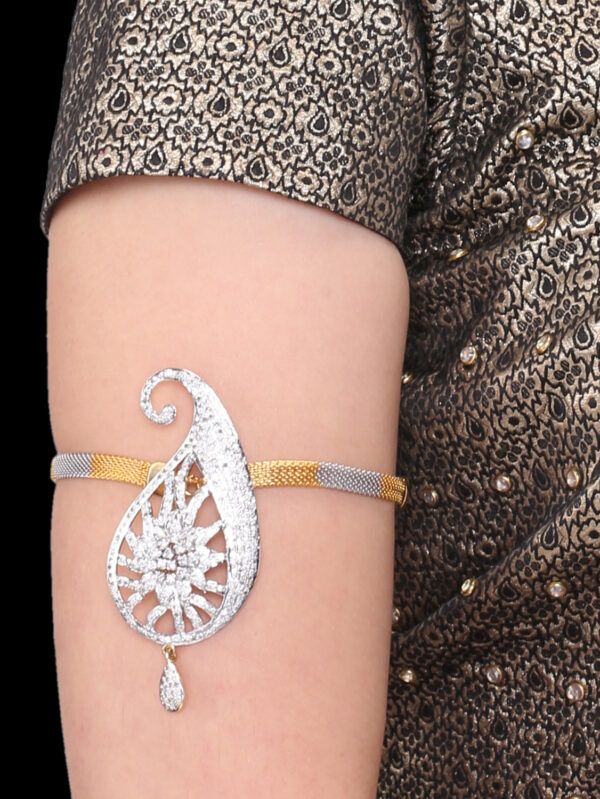 Close-up of a silver teardrop-shaped pendant bracelet with intricate filigree and a dangling teardrop charm on a model's bare upper arm, against a patterned metallic dress.