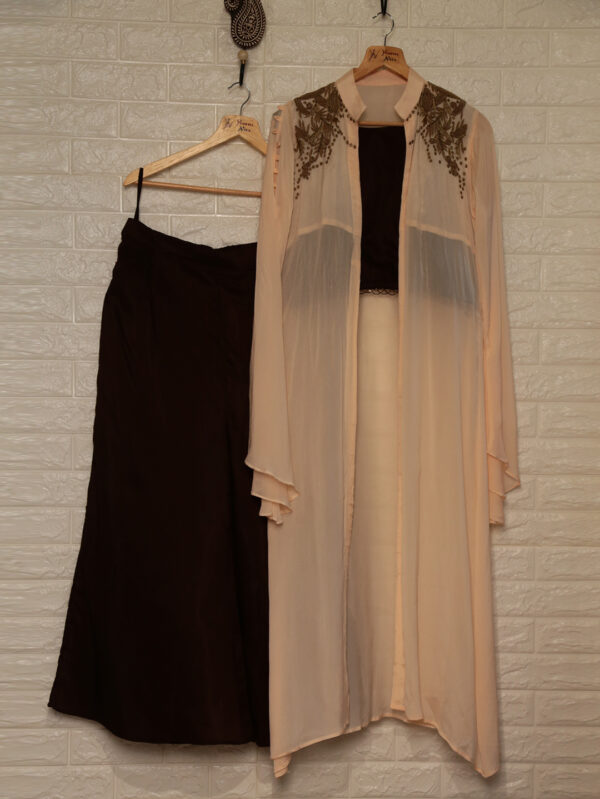 Beige sheer long cardigan with brown embroidery at the shoulders, over a black top, on a hanger beside a dark maxi skirt against a white brick wall.