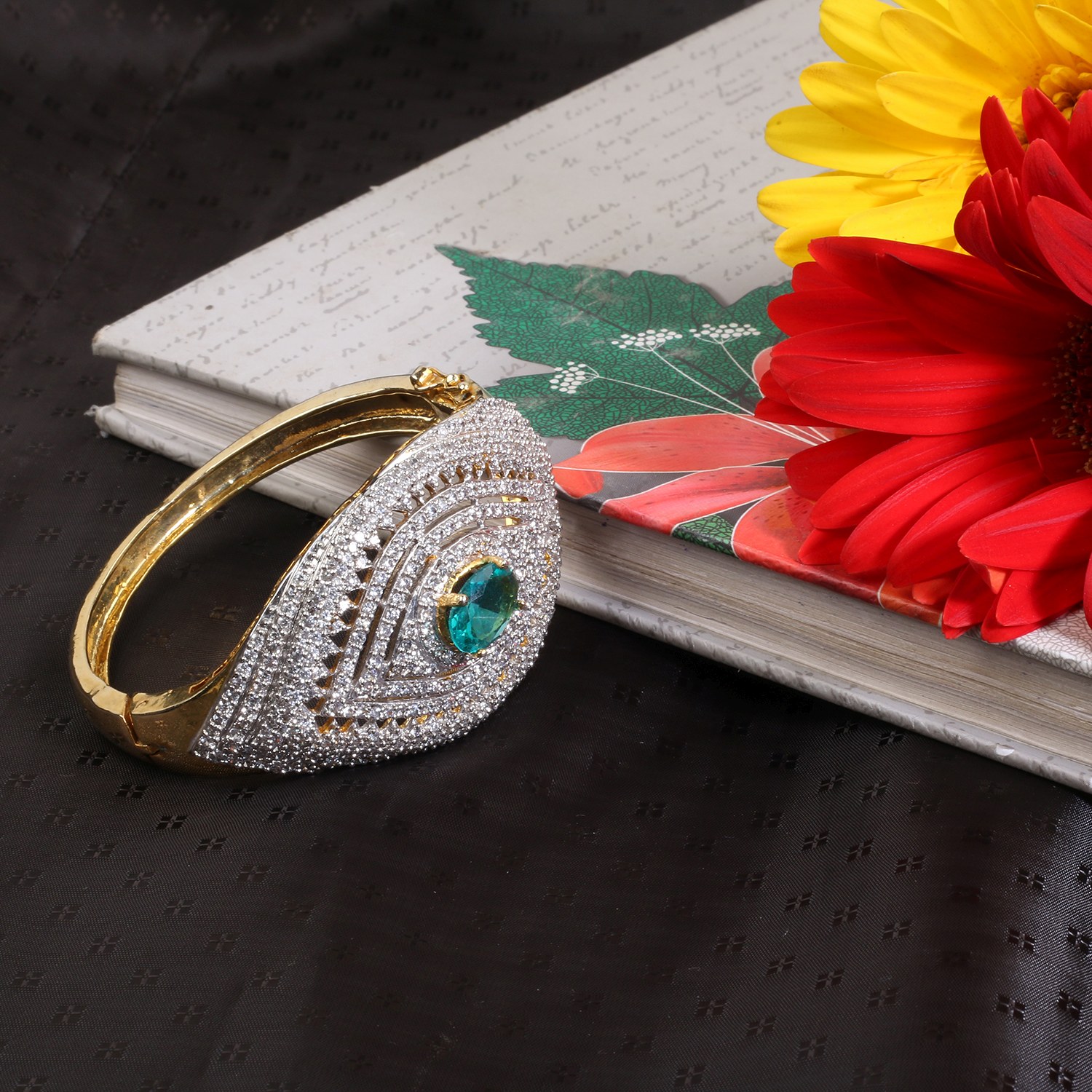 Gold and diamond bracelet with a green gemstone resting on a white notebook beside bright yellow and red flowers at the edge of the page.