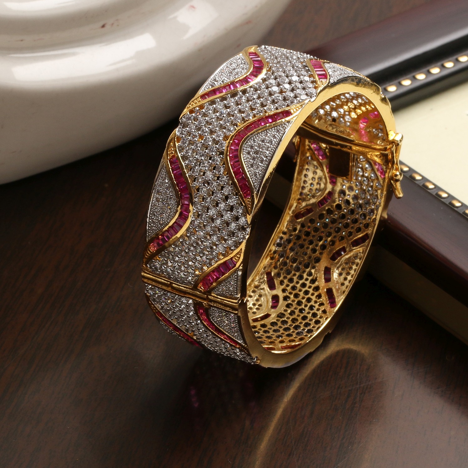 Gold, diamond-studded bangle with pink gemstone inlays resting on a dark wooden surface.