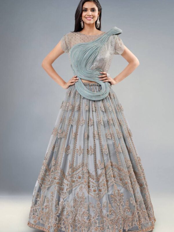 Woman in a light gray, heavily embroidered lehenga with swirling aqua belt and sheer dotted blouse, posing with hands on hips and earrings visible on a neutral backdrop.